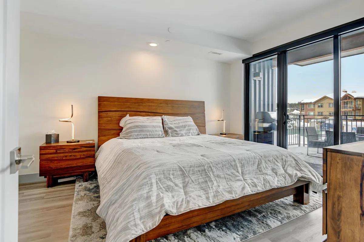 A serene and inviting bedroom featuring a plush bed with premium linens, flanked by nightstands. Natural light pours in from a sliding door that leads to a balcony, offering a tranquil view. The room radiates comfort and privacy, ideal for restful nights after mountain adventures.