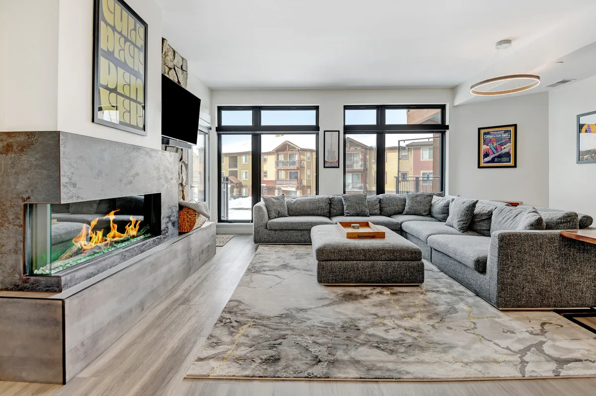 The image showcases a beautifully designed living room featuring a modern fireplace, a spacious sectional couch perfect for lounging, and stylish artwork adorning the walls. Natural light pours in, creating a warm, inviting atmosphere ideal for relaxing after a day on the slopes, socializing with friends and family, or enjoying a quiet evening by the fire.