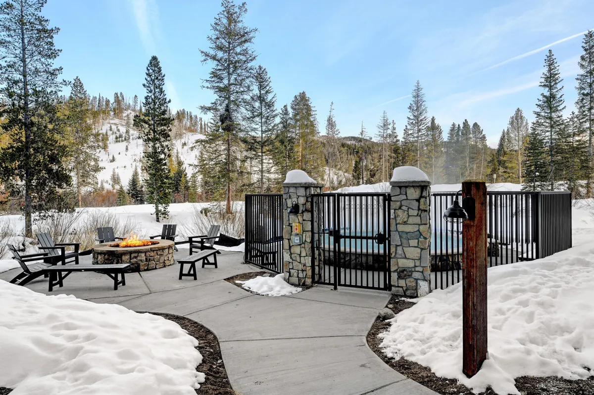 A cozy exterior setup with a fire pit and a hot tub enclosure surrounded by pine trees and a snow-covered landscape. Chairs are placed around the fire, inviting relaxation after a day outdoors.