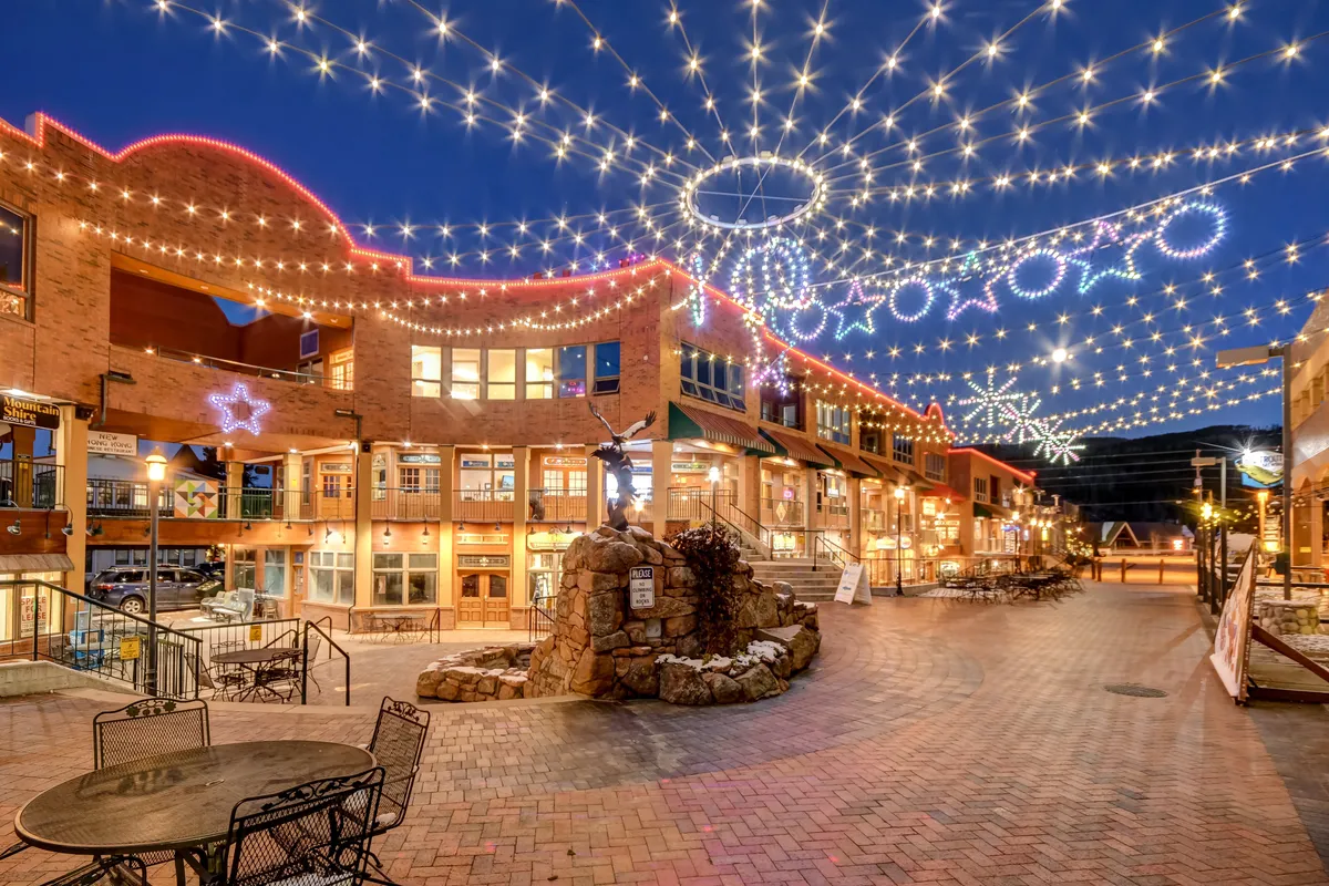 A vibrant outdoor pedestrian area strung with festive lights and decorative star ornaments, featuring a central fountain surrounded by people strolling, shopping, and enjoying al fresco dining. The lively atmosphere is accentuated by evening lighting and visible storefronts.