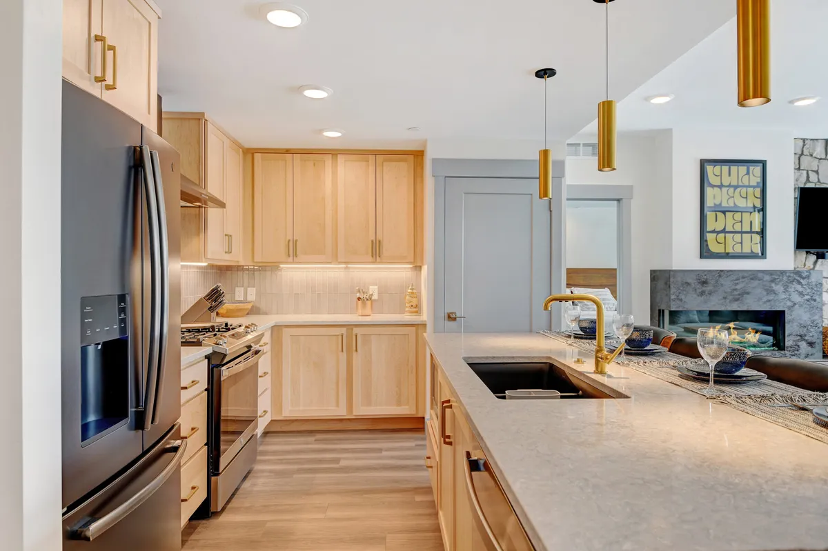 A contemporary kitchen scene featuring a central island with bar stools, stainless steel appliances, gold pendant lights, and sleek natural wood cabinetry. The space is well-lit, modern, and designed for entertaining and group meals.