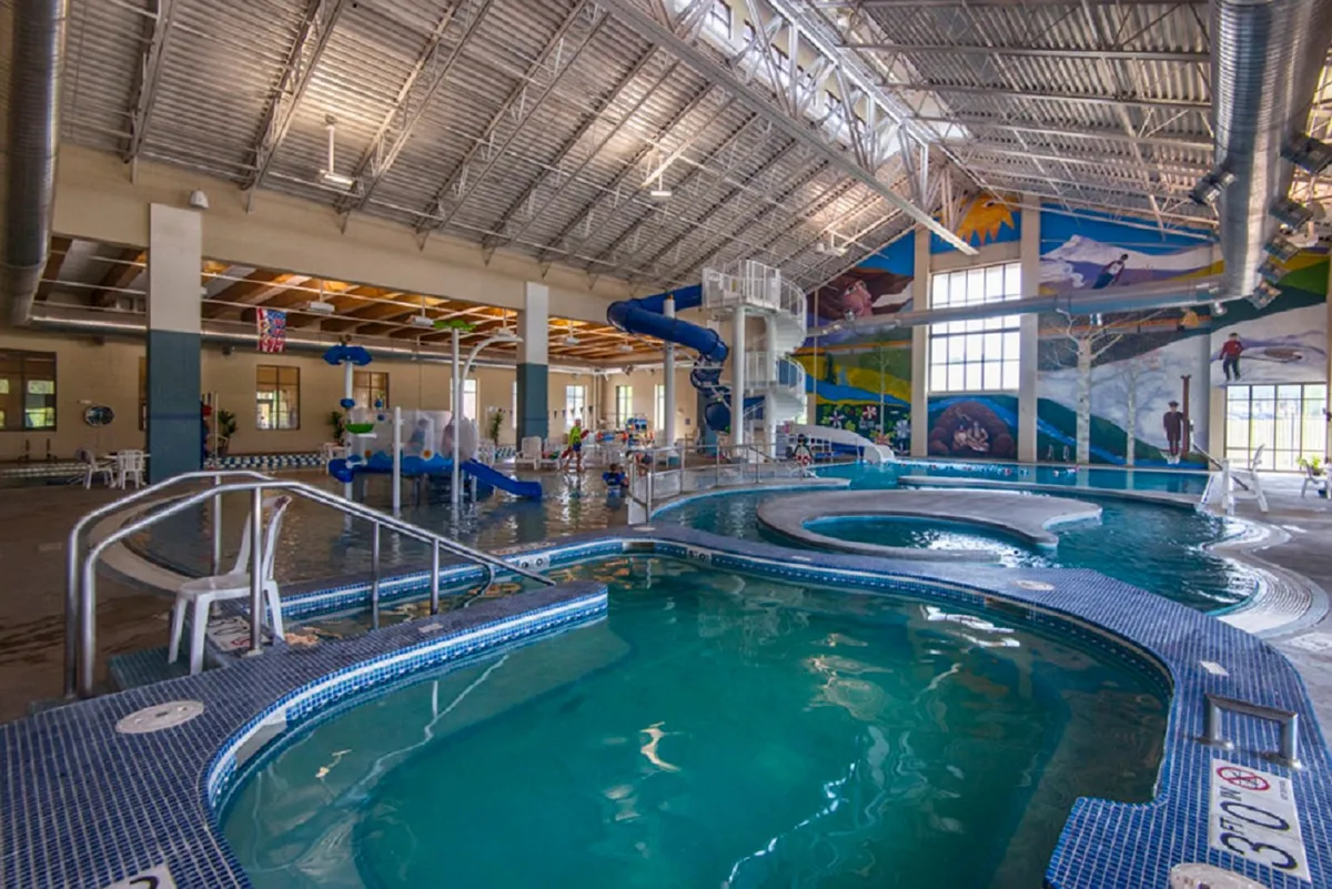 A lively indoor pool area with colorful water slides, a mural, and children playing, capturing the energetic spirit of the nearby Grand Park Rec Center.