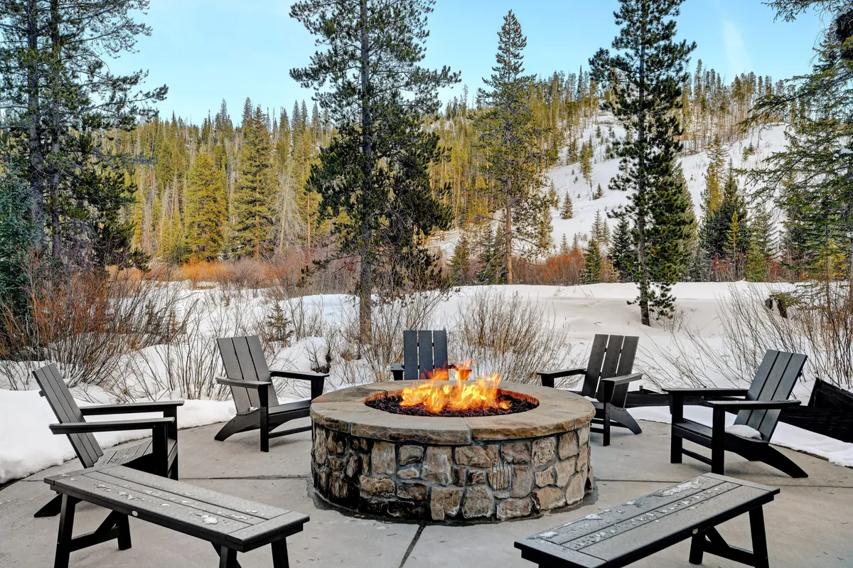 A cozy fire pit surrounded by chairs on snow-covered ground, framed by tall trees and a wintry landscape. The inviting scene suggests warmth, relaxation, and communal moments under the open sky—even in winter.
