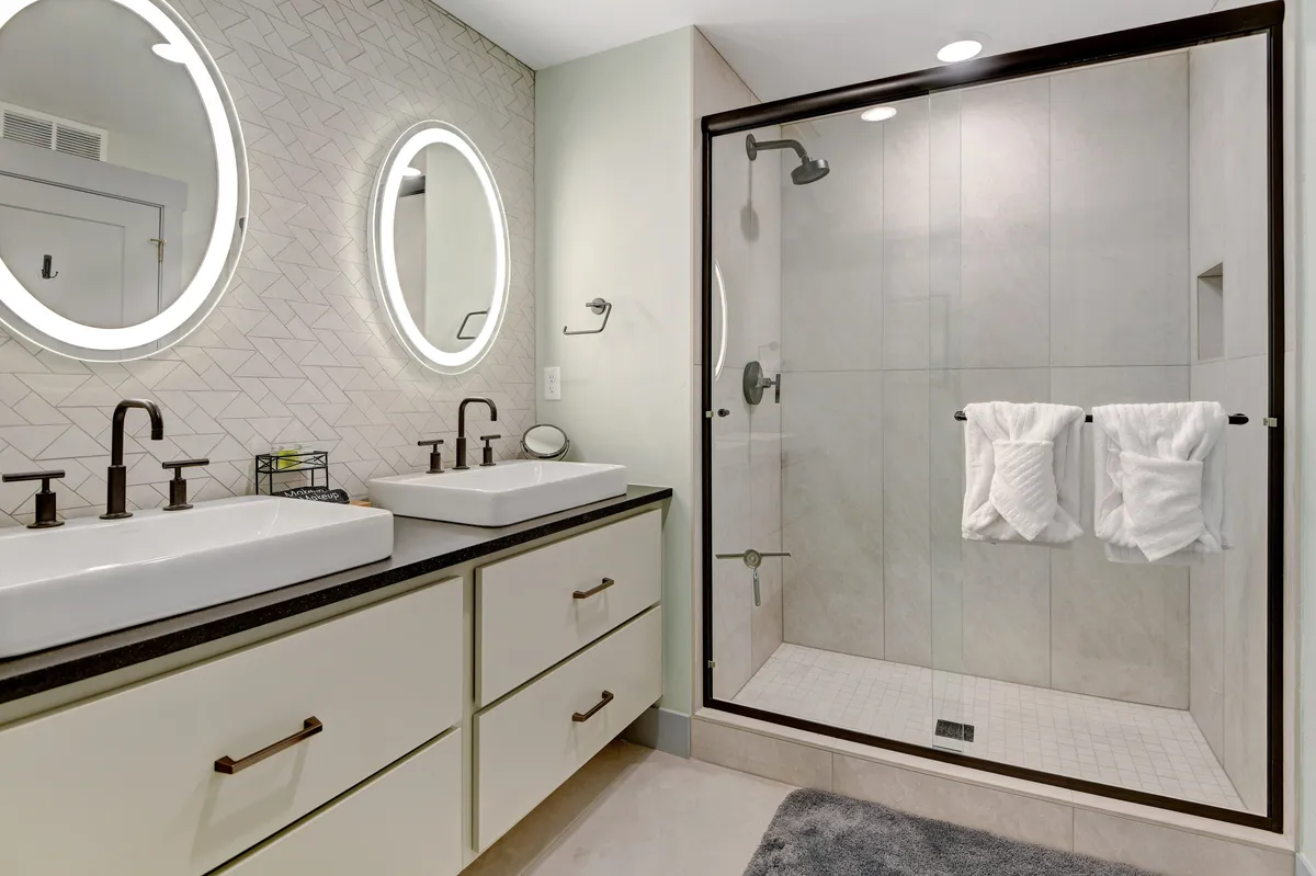 A sophisticated spa-inspired bathroom with double vessel sinks, chic mirrored lighting, and a glass-enclosed walk-in shower. The space is immaculate and luxurious, offering a calming atmosphere for self-care routines or unwinding after a day outdoors.