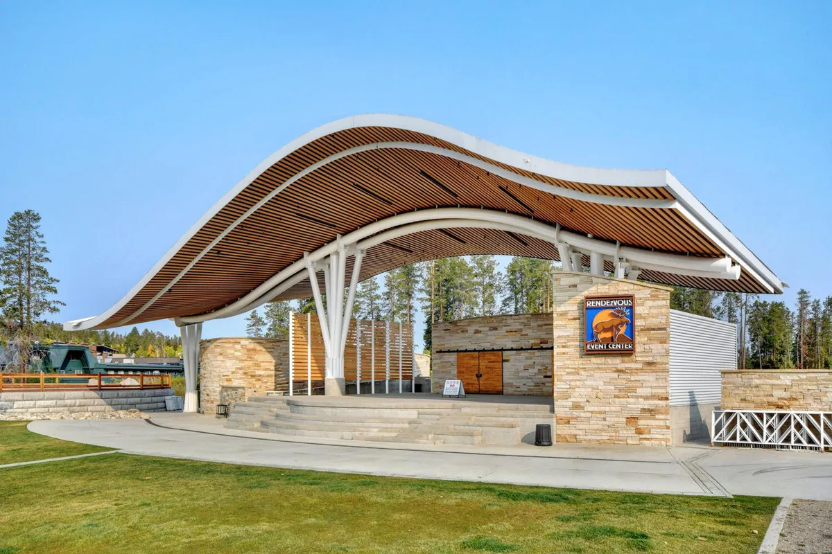 An open-air amphitheater with a unique canopy structure and natural stone walls, set up for live performances and community gatherings. People are seen enjoying a communal, outdoor event, suggesting a lively and engaging environment.