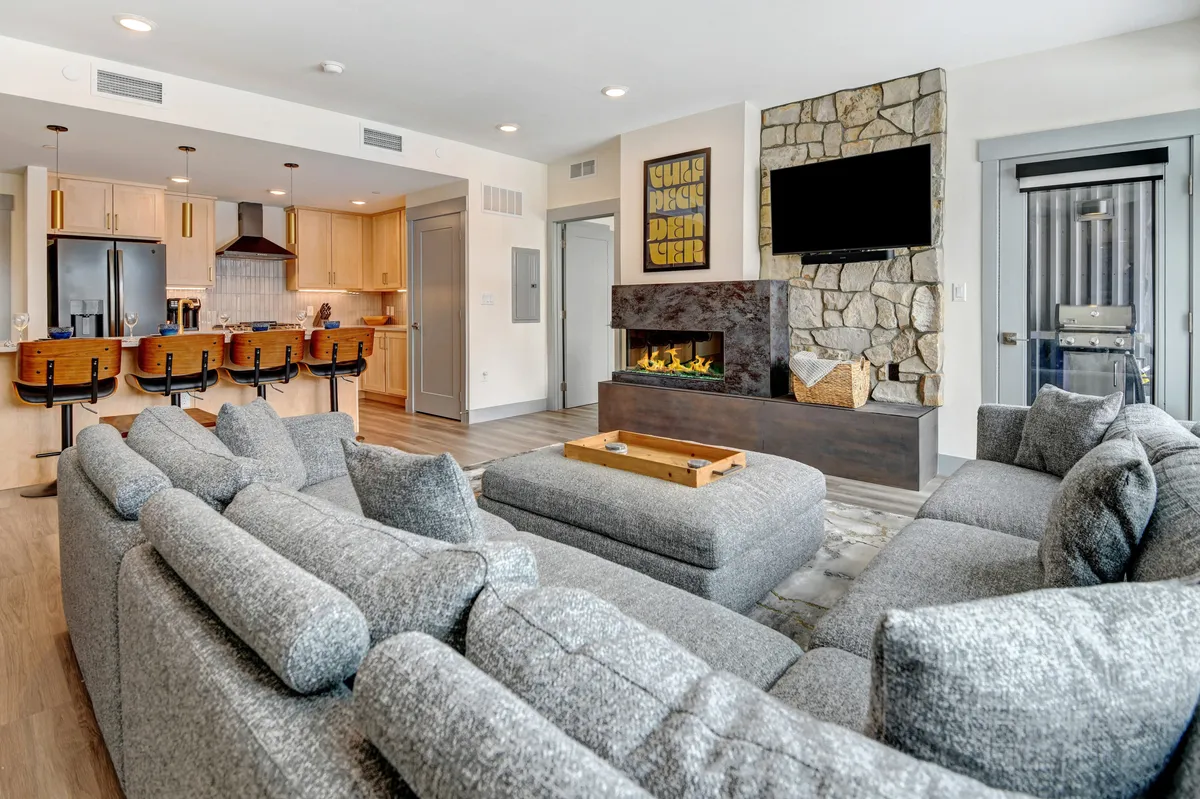 This interior shot captures a spacious open-plan living area with a cozy sectional sofa, a striking color-changing fireplace, a kitchen island with bar seating, and artistic wall décor. The scene is inviting, with plenty of natural light and a seamless flow between the living, dining, and kitchen spaces.